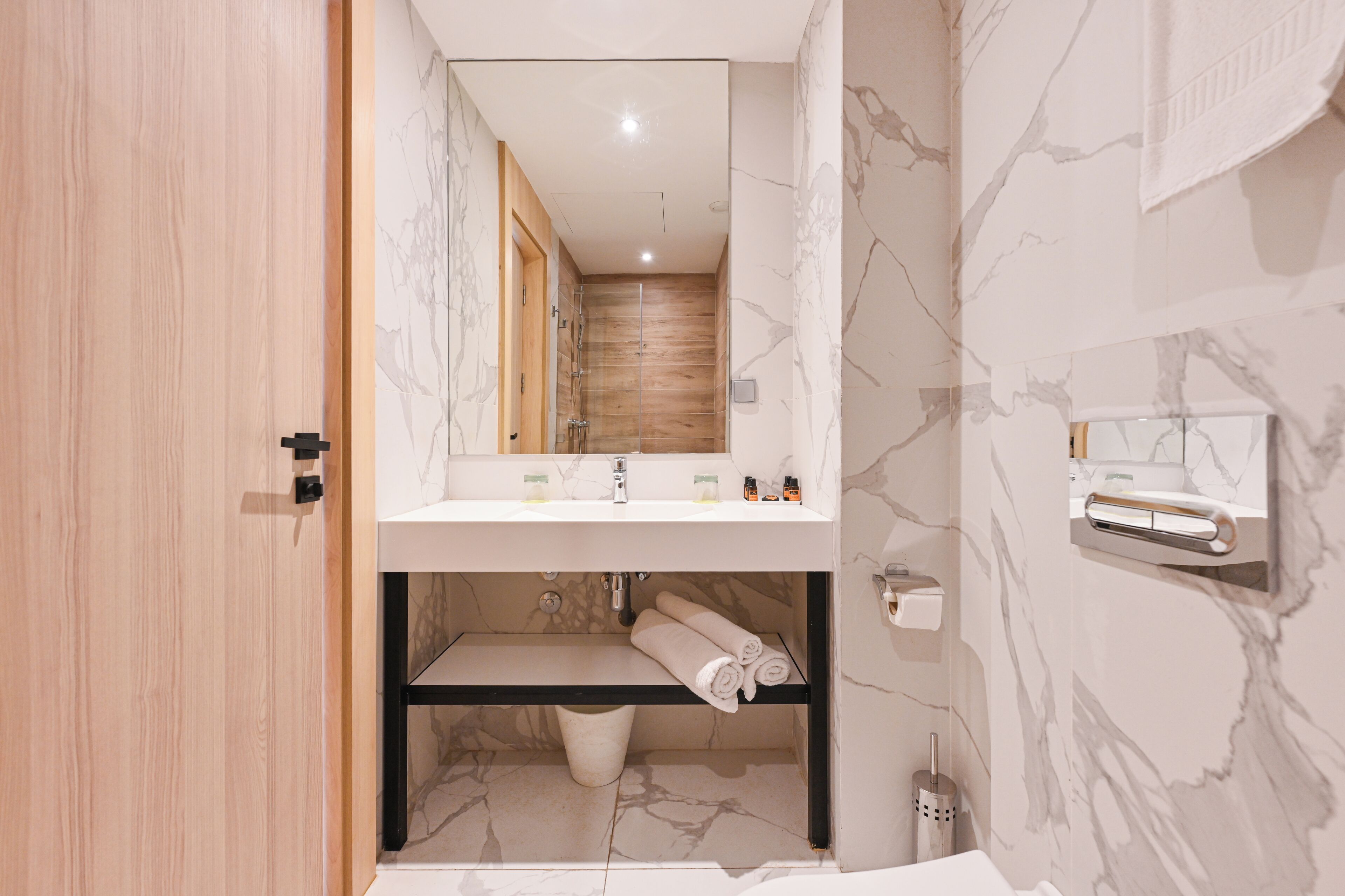 Un baño moderno con puerta de madera, pared de mármol, lavabo con espejo, toallero y zona de ducha.