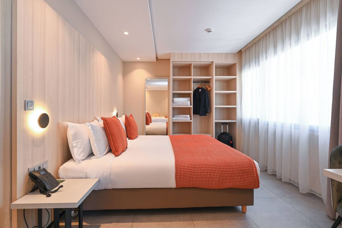 A modern hotel room with a large bed, a bedside table with a phone, a wardrobe, and a mirror.