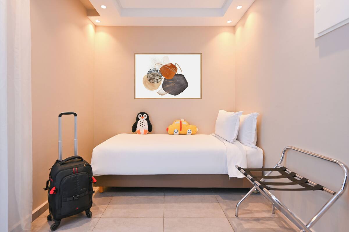 A hotel room with a bed, a framed artwork on the wall, a luggage trolley, and a bench.