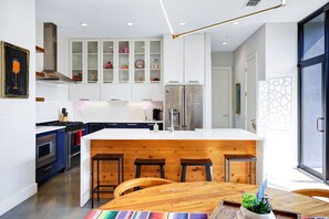 A modern kitchen with white cabinetry, a wooden island with bar stools, stainless steel appliances, and a large window providing natural light. - Private Southtown Compound | Rooftop Terrace | Walk to Alamodome & River Walk (San Antonio)