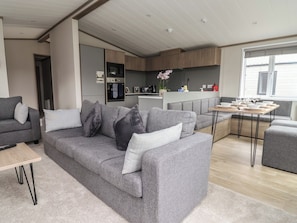 A modern living room with a grey sofa, a small wooden table, and a kitchen in the background. - Cedar Lodge 2 Bedrooms With hot tub (Northwich)