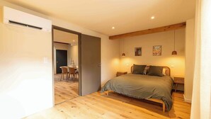 A bedroom with a wooden floor, a bed with a grey bedspread, two bedside lamps, and a small table with chairs in the adjacent room. - Apartment "Origan" (Saint-Rémy-de-Provence)