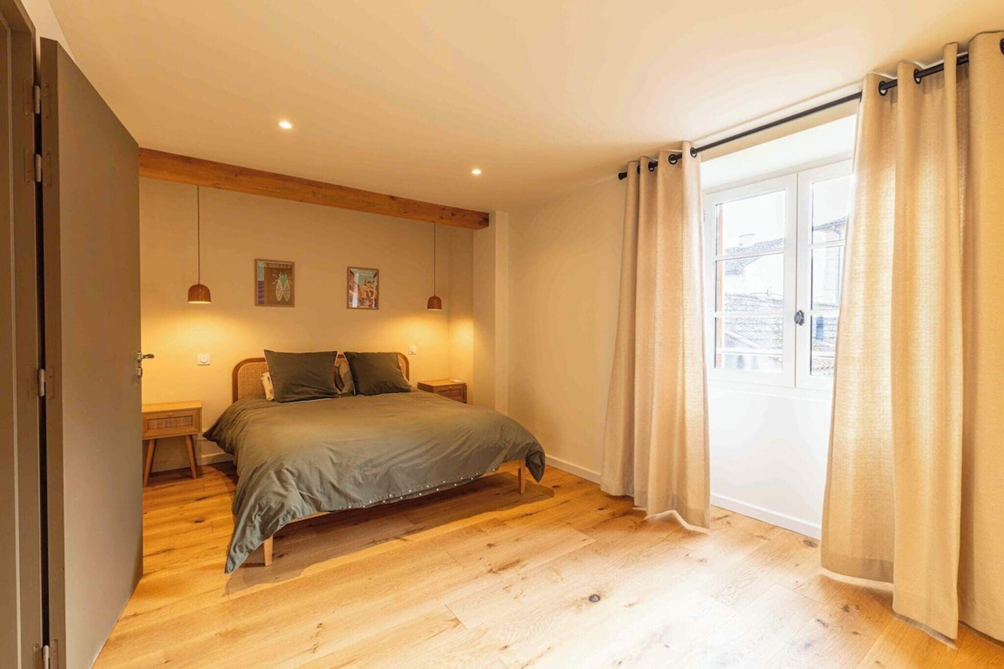 Ein Schlafzimmer mit Holzboden, einem Bett mit grauer Bettdecke, zwei Nachttischen, einem Fenster mit Vorhängen und Blick auf Gebäude.