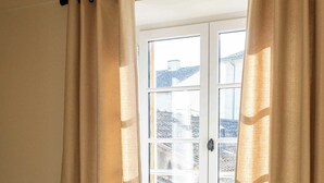 A room with a wooden floor, a window with curtains, and a view of buildings outside. - Apartment "Origan" (Saint-Rémy-de-Provence)