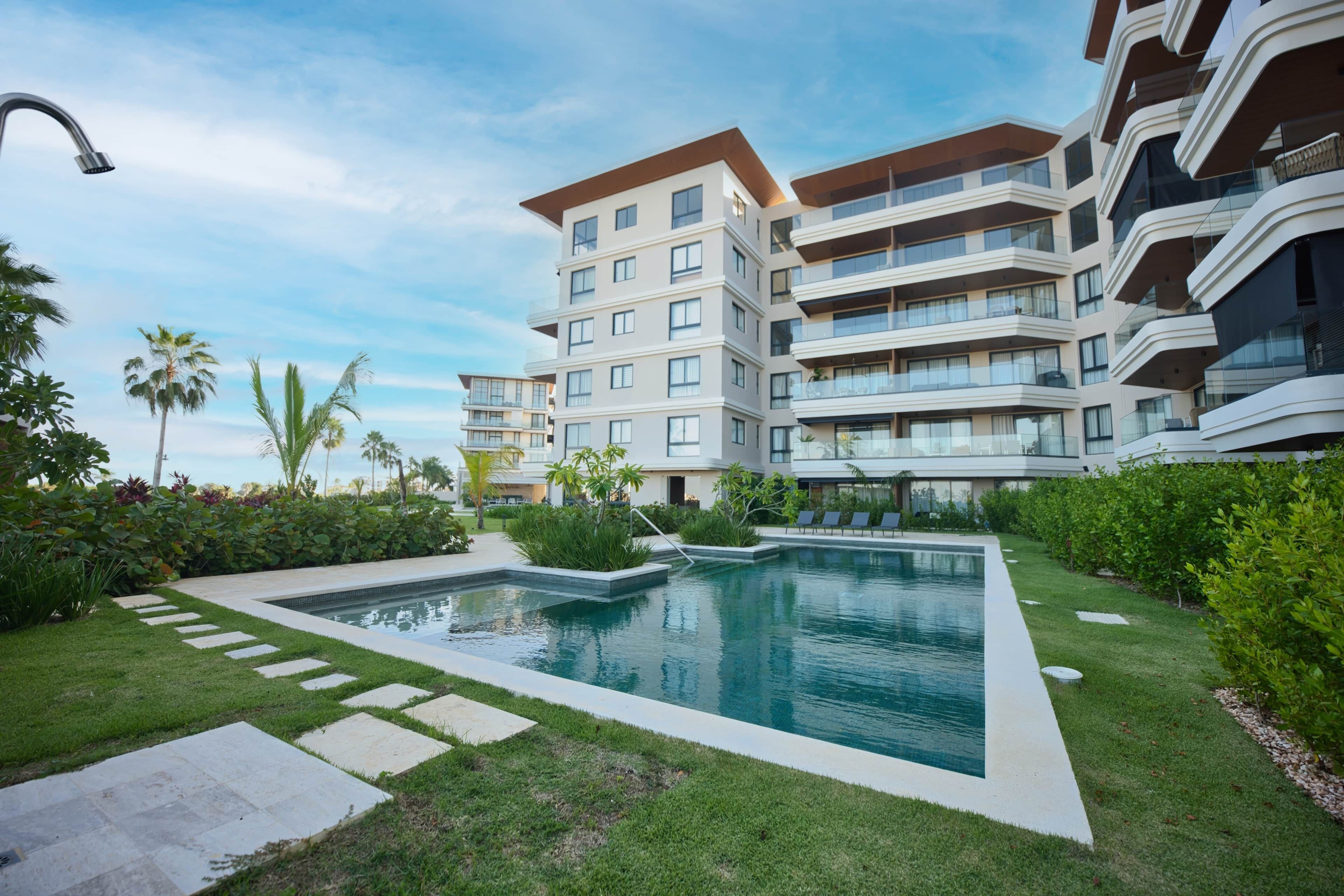 Un edifici residencial modern amb piscina, envoltat de gespa ben cuidada i palmeres.
