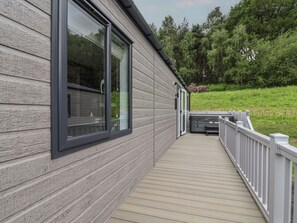 A wooden deck with a white railing, a large window, and a view of a green landscape. - Cedar Lodge 3 Bedrooms With hot tub (Northwich)