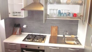 A compact kitchen with a gas stove, a sink, and a countertop. There are cabinets below the countertop and a shelf above it holding dishes and glassware. - Superb apartment with sea view, right in the center of town (Antsiranana)