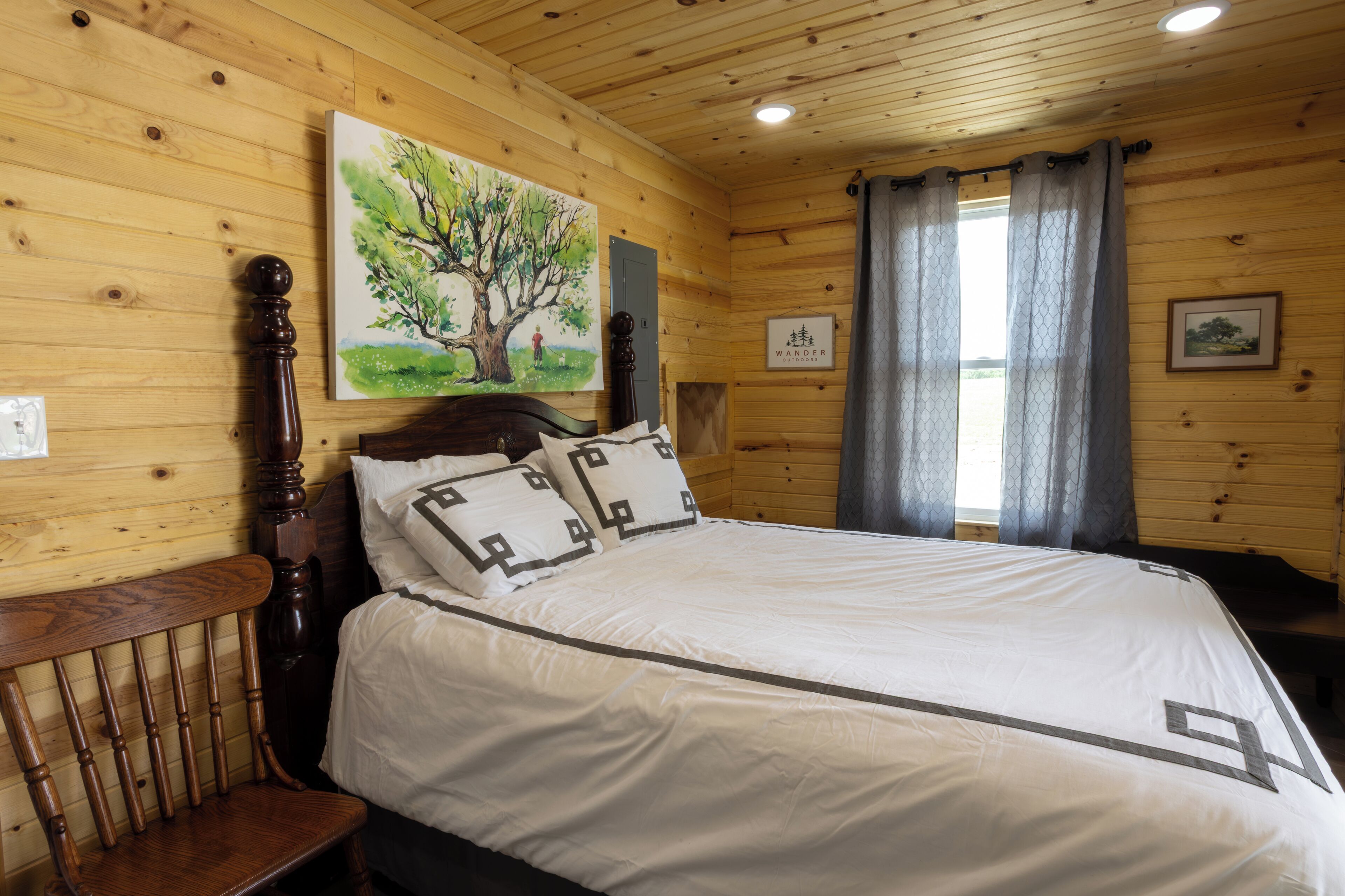 A wooden cabin room with a bed, a chair, a painting of a tree, and a window with curtains.