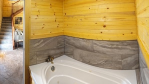Interior de una cabaña de madera con una bañera grande, una escalera de madera y suelo de baldosas.