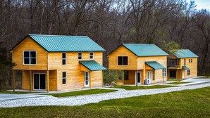 Una hilera de cabañas de madera con techos verdes, cada una con puerta principal y ventanas, con un bosque de fondo.