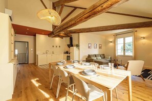 Un'area pranzo con tavolo in legno apparecchiato per quattro persone, una cucina sullo sfondo e un soggiorno con divano e finestra.