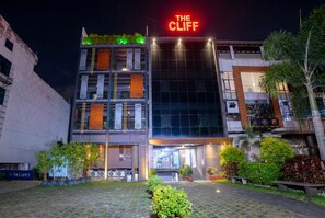 Un hôtel à la façade moderne, avec une enseigne lumineuse au néon indiquant « THE CLIFF », et une cour dallée agrémentée de plantes en pot.