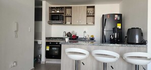 A modern kitchen with a built-in microwave, a refrigerator, a coffee maker, and a stove. There are three white bar stools in front of the counter. - Mirador Baluarte Pool& Parking (Cartagena de Indias)