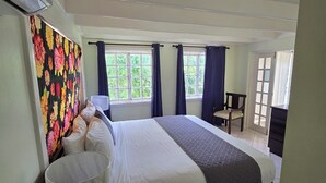 A bedroom with a large bed, a bedside table, a chair, a window with curtains, and a view of greenery outside. - Live Easy at the mystic (Ocho Rios)