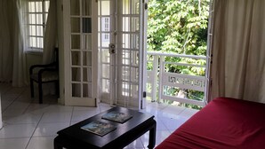 A room with a red bed, a black table with two books, a chair, and a balcony with a white railing and greenery outside. - Live Easy at the mystic (Ocho Rios)
