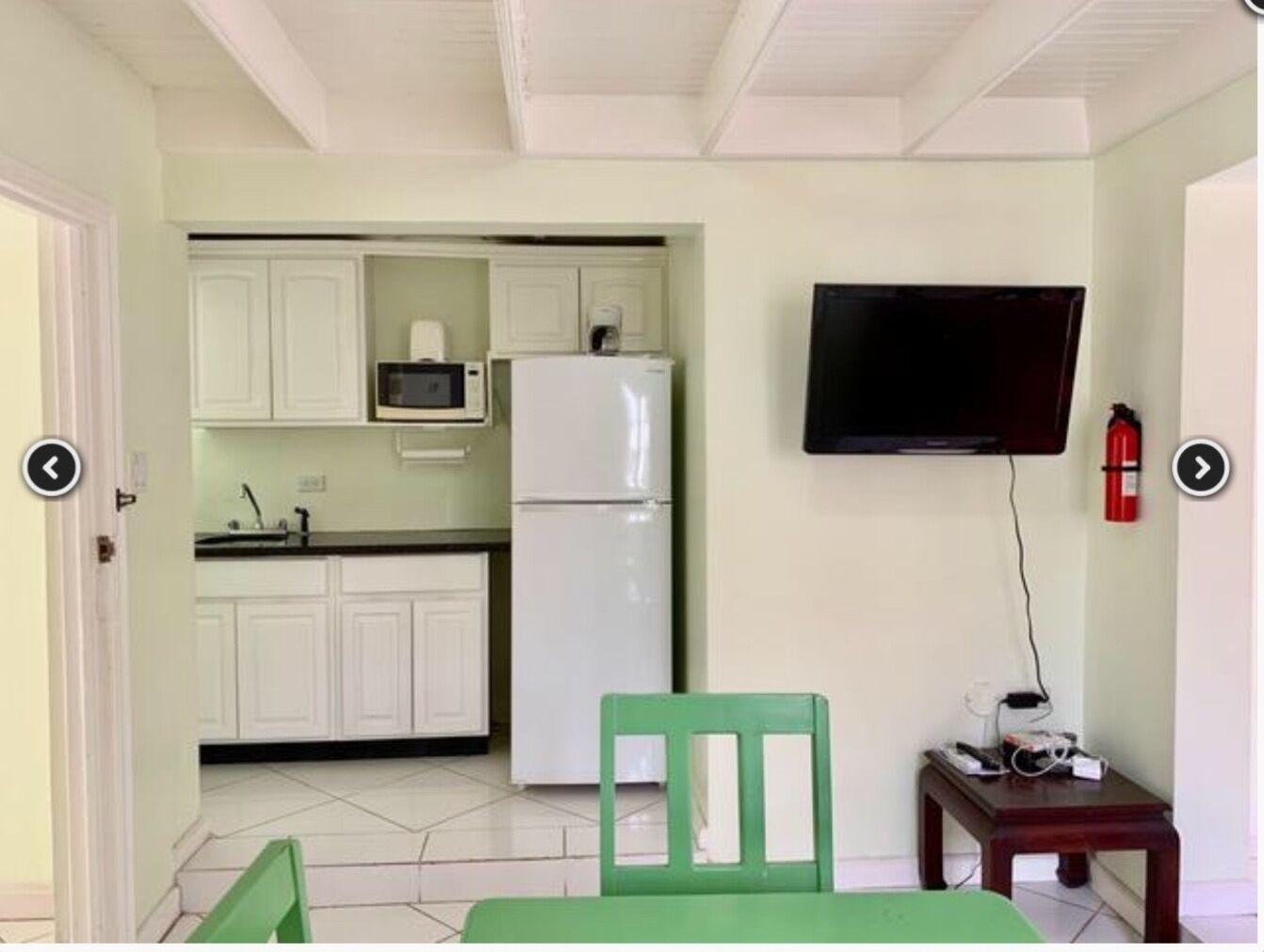 Une cuisine compacte avec des meubles blancs, un four à micro-ondes et un réfrigérateur. On y trouve une chaise verte et une petite table, ainsi qu’un téléviseur fixé au mur.