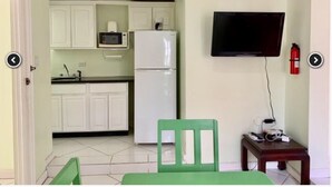 A compact kitchen with white cabinets, a microwave, and a refrigerator. There is a green chair and a small table with a TV mounted on the wall. - Live Easy at the mystic (Ocho Rios)