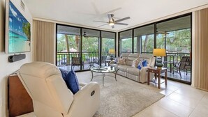 Ein großzügiger Wohnbereich mit Sofa, Sessel und Couchtisch, große Fenster mit Blick ins Freie und ein Deckenventilator.