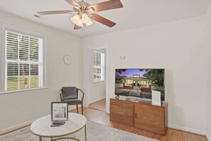 A room with a ceiling fan, a television displaying a house, a chair, a small table with a keyboard, and a wooden cabinet. - Charming Charleston Bungalow Near Park Circle (North Charleston)