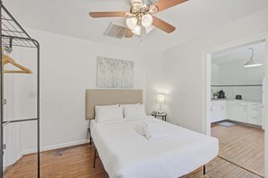 A hotel room with a bed, a ceiling fan, a painting, a nightstand, and a view of the kitchen. - Charming Charleston Bungalow Near Park Circle (North Charleston)