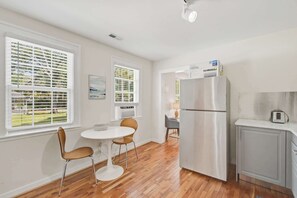 A modern kitchen with a stainless steel refrigerator, a small dining area with two chairs, and a window with blinds. - Charming Charleston Bungalow Near Park Circle (North Charleston)