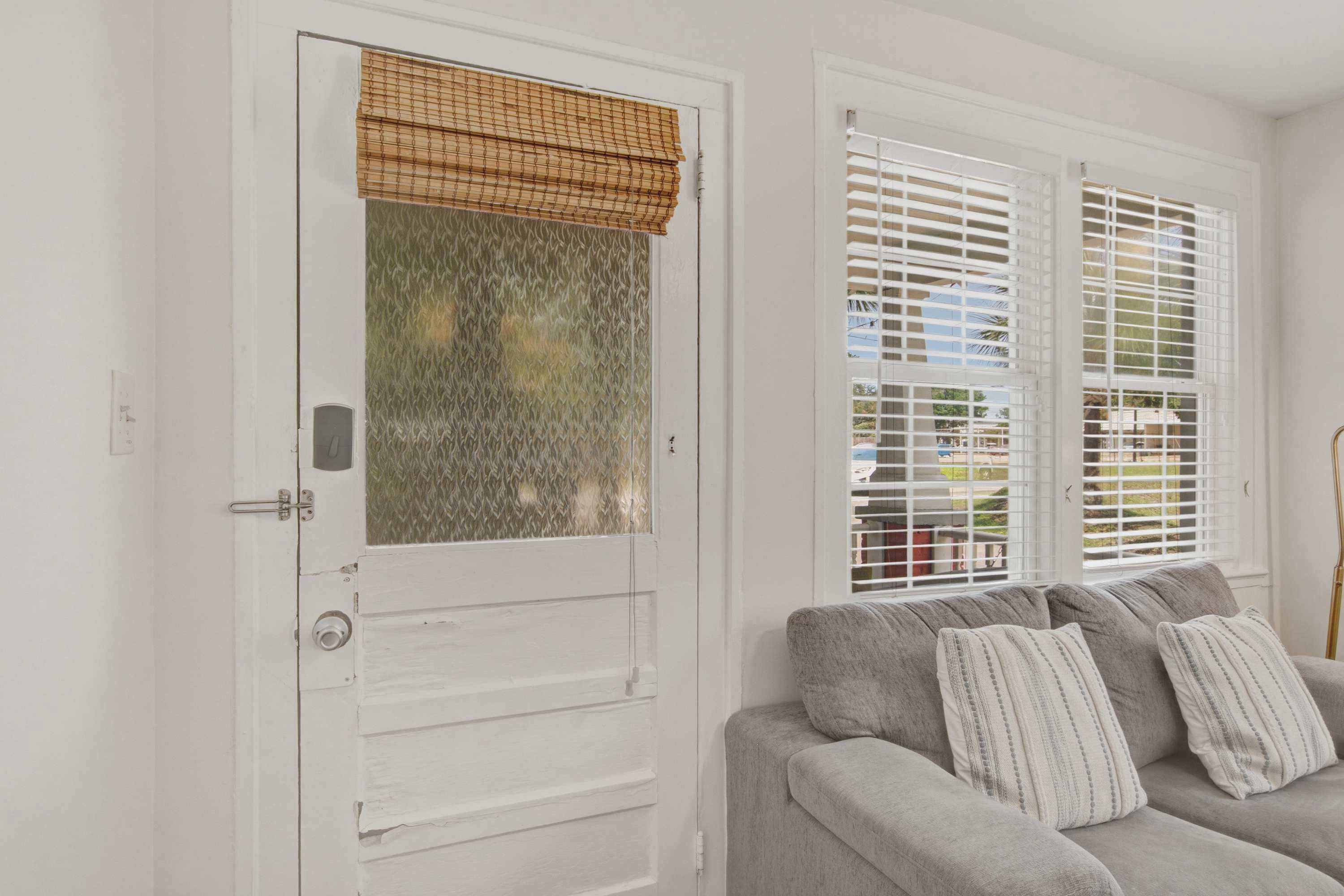 A room with a white door, a window with blinds, and a grey sofa with pillows.