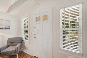 A room with a white door, a window with blinds, a gray sofa, and a framed picture on the wall. - Stylish Bungalow Stay Near Downtown Charleston (North Charleston)