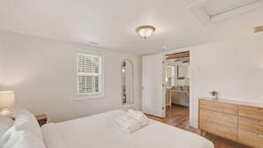 Een slaapkamer met een bed, nachtkastje, lamp, raam met zonwering, spiegel, dressoir en een open deur naar een andere kamer.