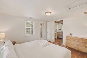 A bedroom with a bed, bedside table, lamp, window with blinds, mirror, dresser, and an open door leading to another room. - Stylish Bungalow Stay Near Downtown Charleston (North Charleston)