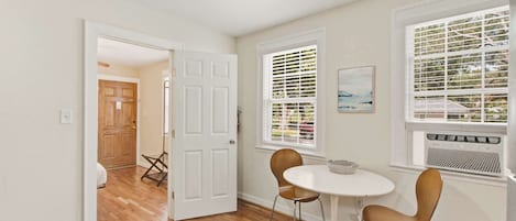 Ein Zimmer mit Holzboden, weißen Wänden, einer weißen Tür und einem Fenster mit Jalousien. Ein Tisch mit zwei Stühlen und ein kleines Gemälde sind an der Wand angebracht.