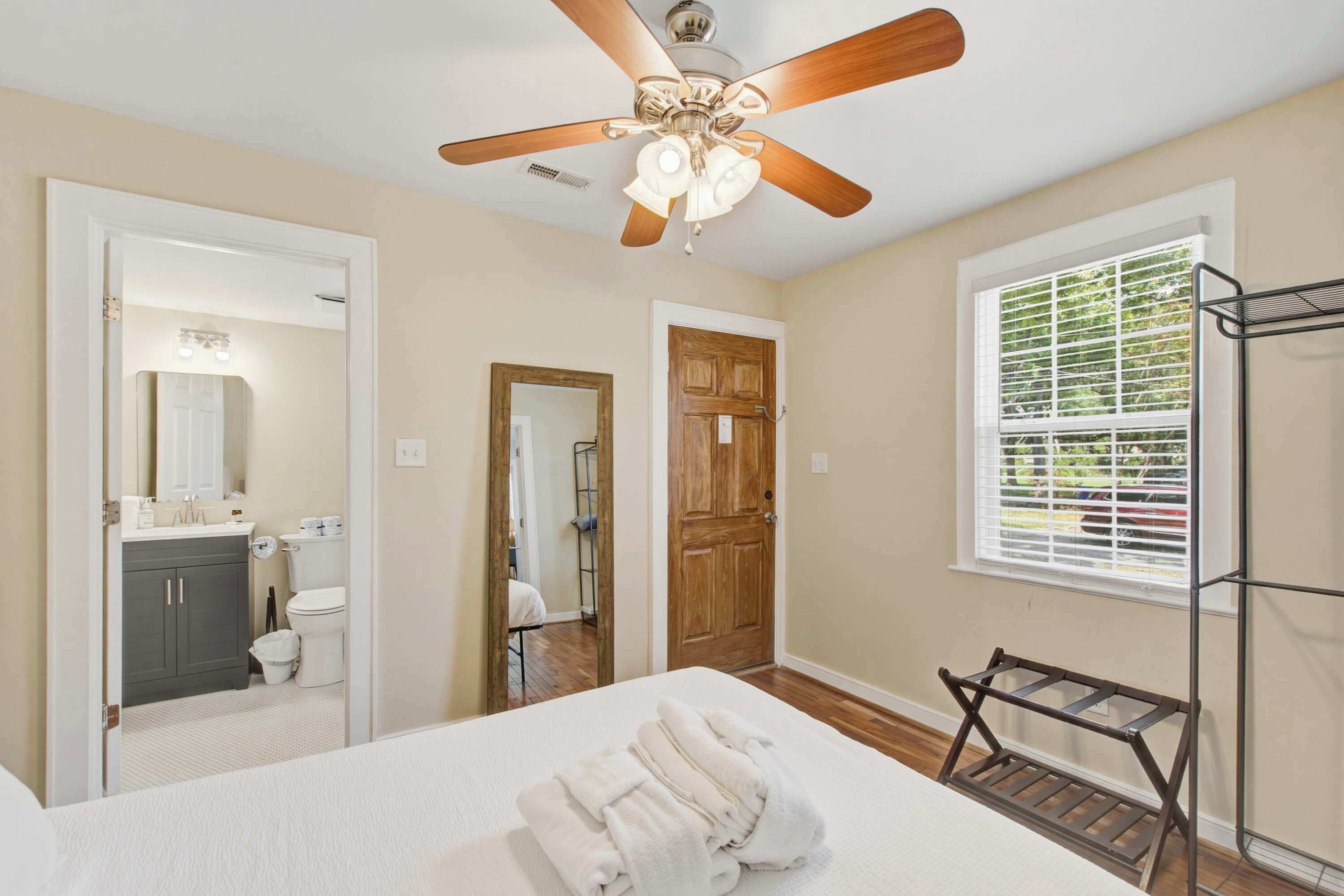 A hotel room with a ceiling fan, a bed with white bedding, a wooden door, a window with blinds, and a metal rack.