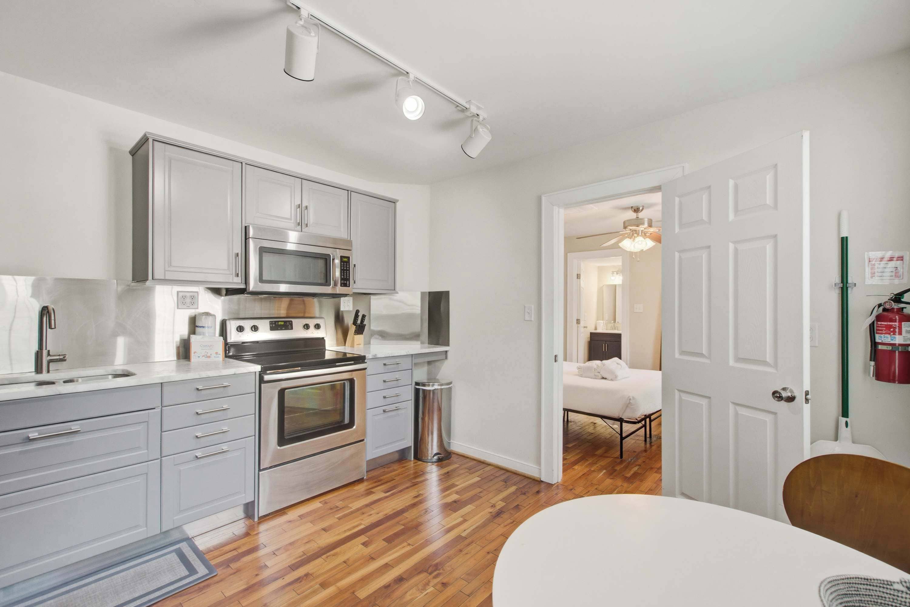 A modern kitchen with stainless steel appliances, a dining area with a white table, and a visible hallway leading to a bedroom.