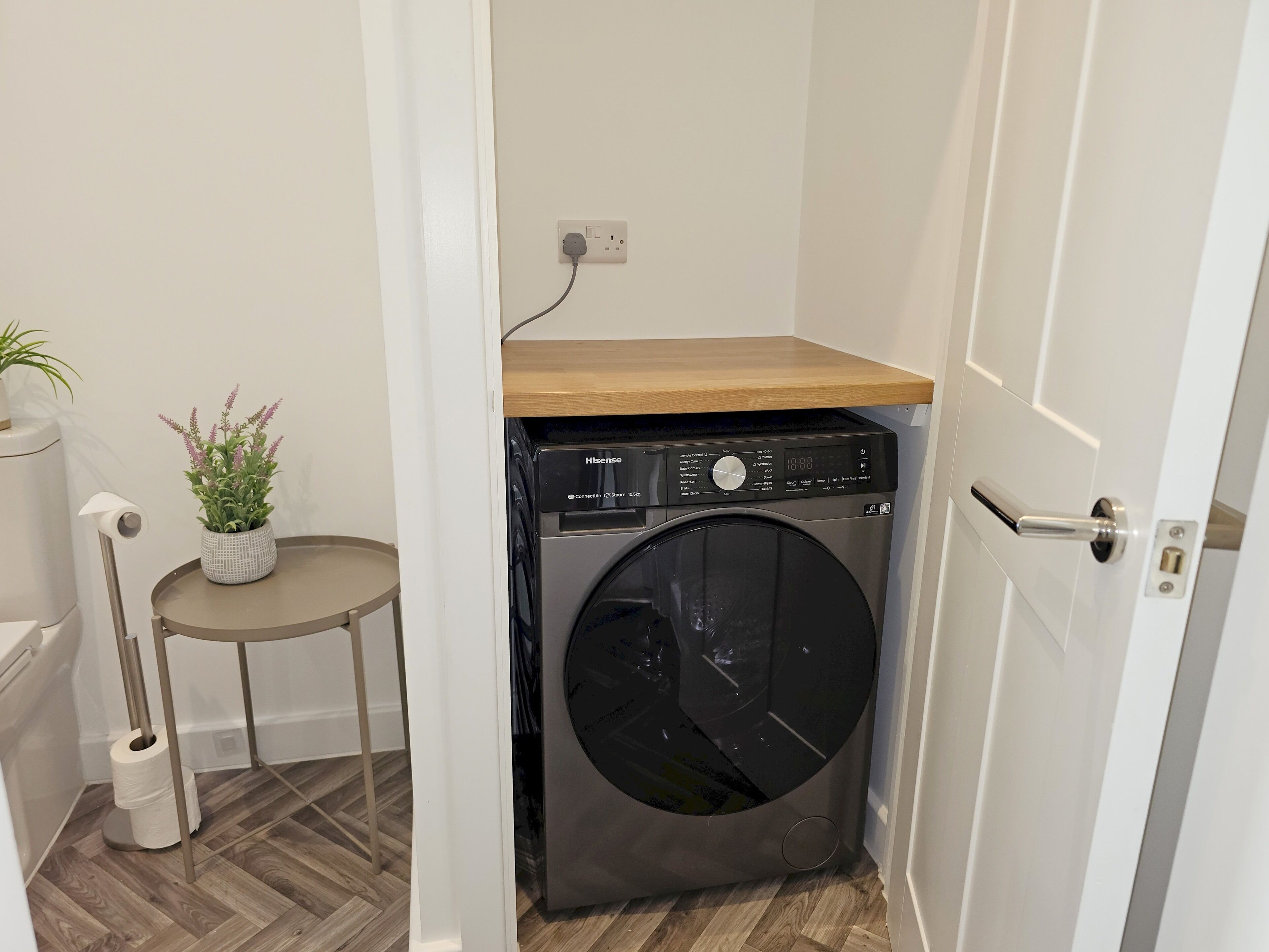 Une petite buanderie comprenant un lave-linge, une petite table avec un pot de fleurs et un porte-rouleau de papier toilette.