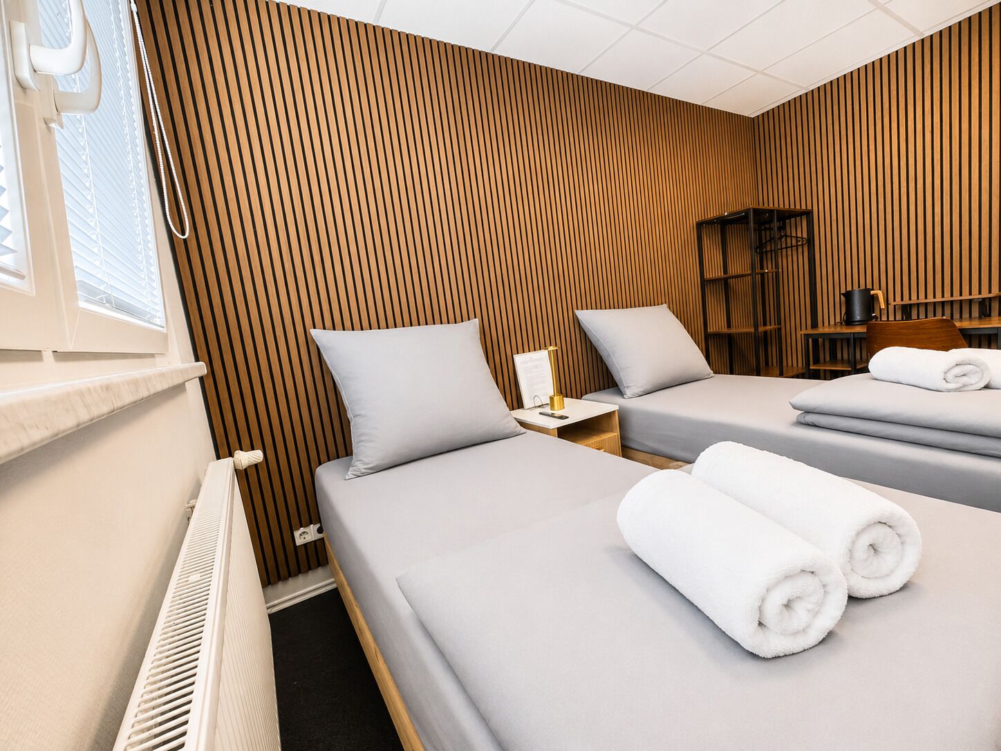 A hotel room with two beds, a radiator, a nightstand with a lamp, and a bookshelf.