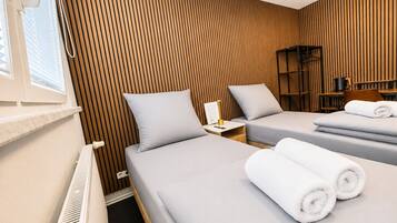 A hotel room with two beds, a radiator, a nightstand with a lamp, and a bookshelf.