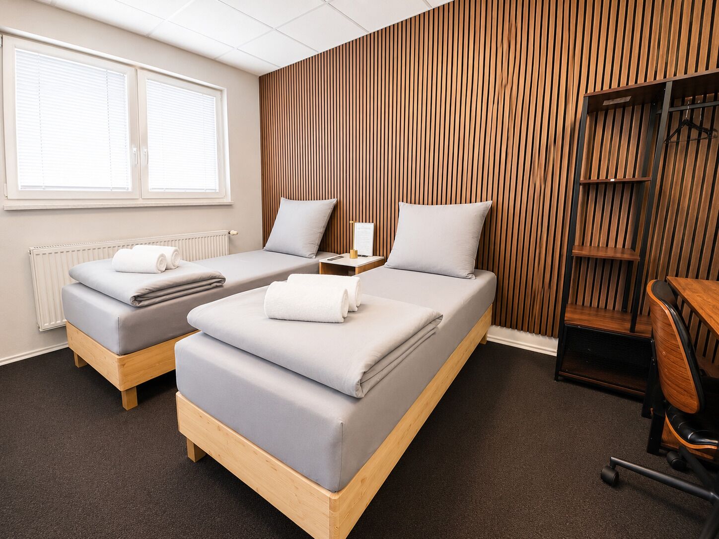 A hotel room with two beds, a desk, a chair, and a radiator.