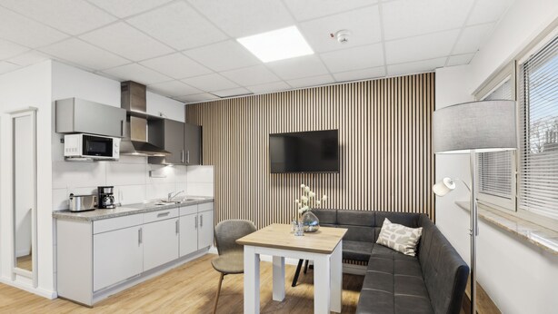 A modern kitchen with white cabinets, a microwave, and a stove. There is a dining area with a table and chairs, and a gray sofa with a TV above it. - HanseStay Wandsbek (Hamburg)