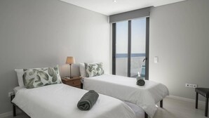 Una habitación de hotel moderna con dos camas, una mesita de noche, una lámpara y vista al mar a través de un gran ventanal corredero.