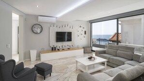 Una sala de estar moderna con un televisor grande, sofás grises, una mesa de centro y vista al mar.