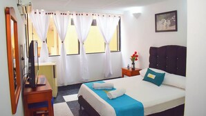 A hotel room with a bed, a desk, a chair, a TV, and a window with curtains. - Hotel Dinastia Real (Neiva)