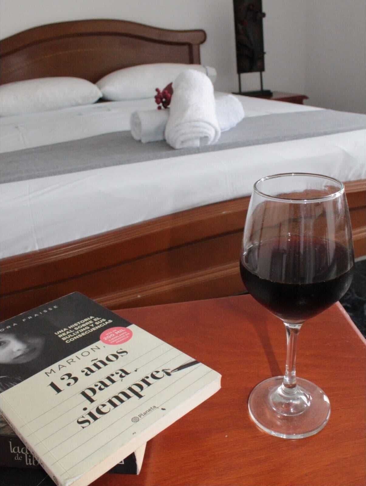 Una camera d'albergo con un letto rifatto, un bicchiere di vino rosso e un libro sul tavolo.