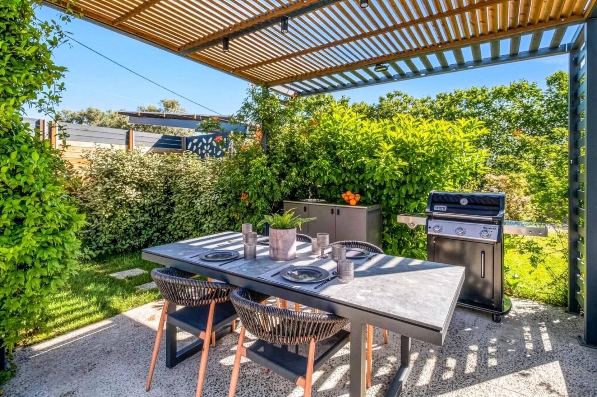 Eine Terrasse im Freien mit Esstisch, umgeben von Grünpflanzen und einer Grillstation.