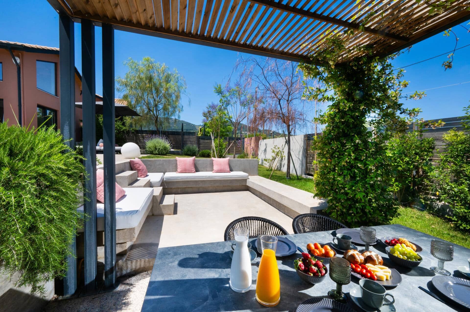 An outdoor patio with a dining table set for a meal, surrounded by greenery and a modern outdoor seating area.