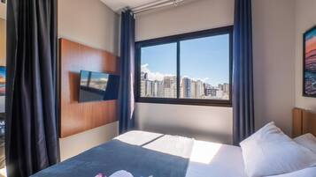 A hotel room with a bed, a TV mounted on the wall, a window with a city view, and curtains.