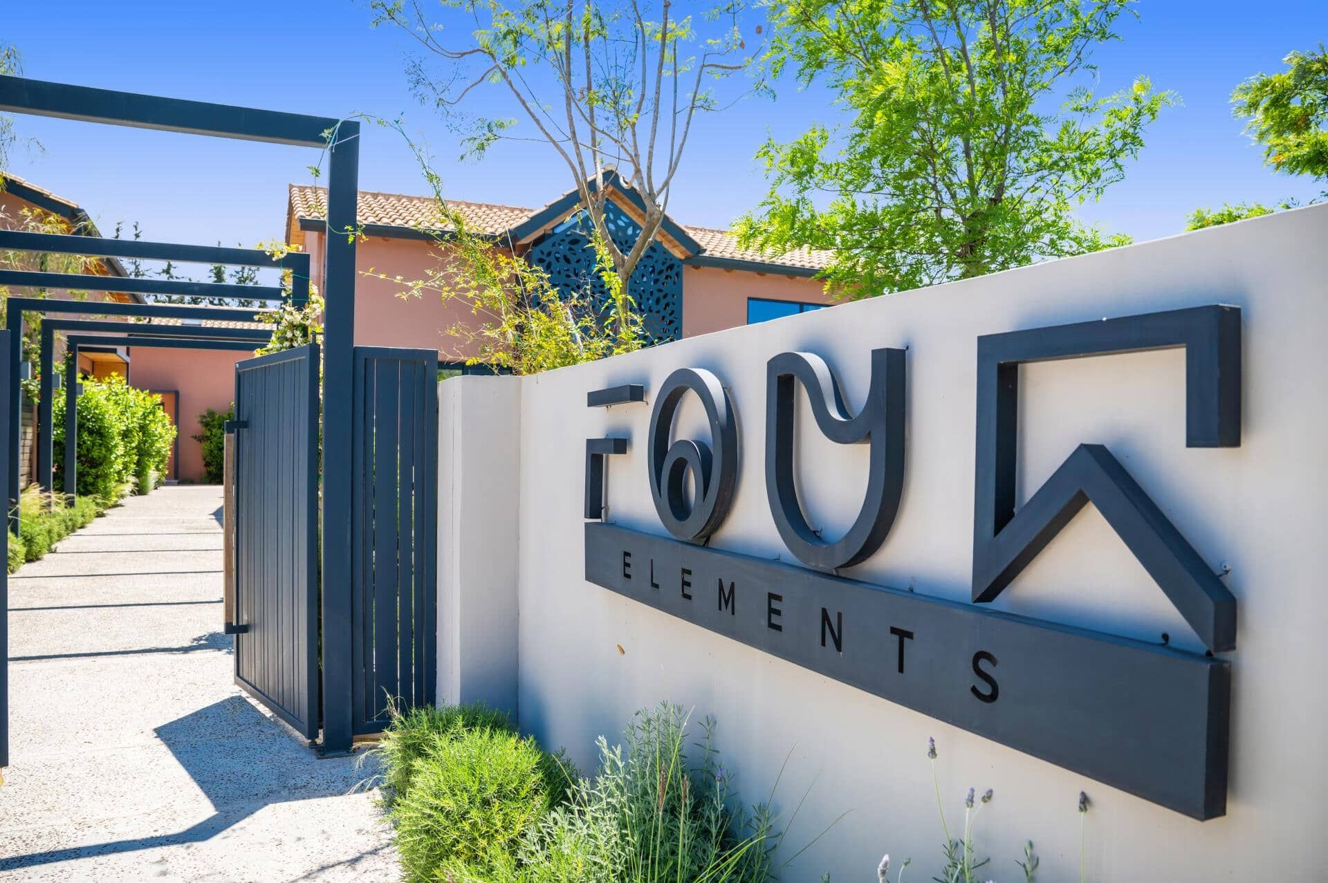 Ein Schild mit der Aufschrift 'FOU ELEMENTS' in modernem Design, ein verschlossenes Tor und ein Weg zu einem Gebäude.