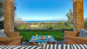 Un patio al aire libre con mobiliario de mimbre, una mesa con un libro y un vaso de jugo de naranja, y una vista panorámica del mar y las montañas.