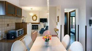 Un comedor moderno con una mesa blanca, sillas y un jarrón de flores. Cuenta con una zona de cocina con armarios y un microondas.