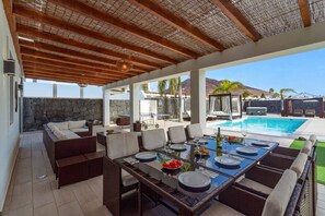A covered outdoor dining area with a pool, wicker furniture, and a stone wall. - Villa Yanira by Villa Plus (Montaña Roja)