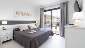 Ein modernes Schlafzimmer mit einem großen Bett, einem Nachttisch, einem Schreibtisch mit Lampe und Blick ins Freie.