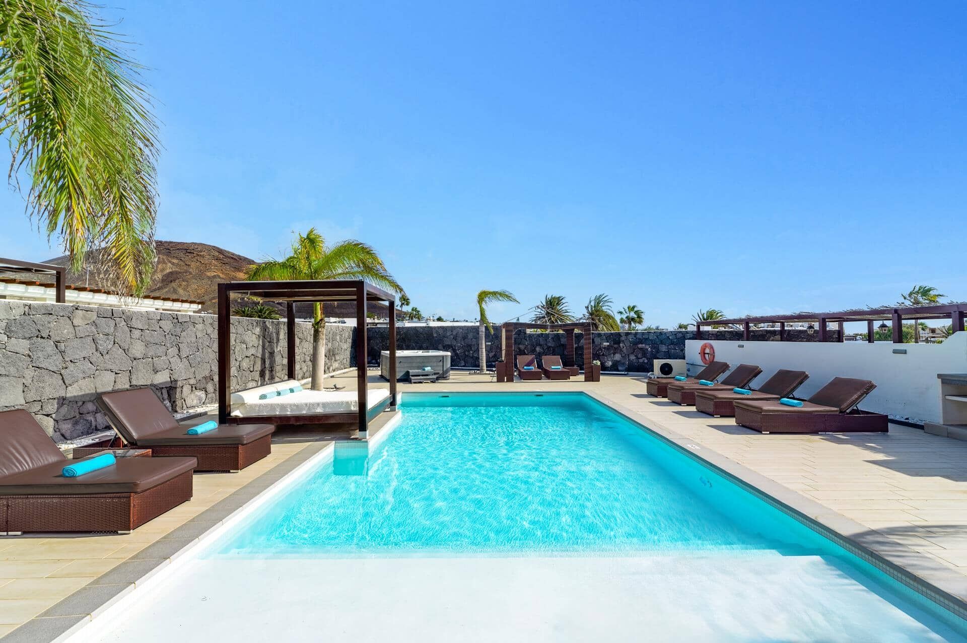 A swimming pool with lounge chairs and a covered seating area, surrounded by stone walls and palm trees.